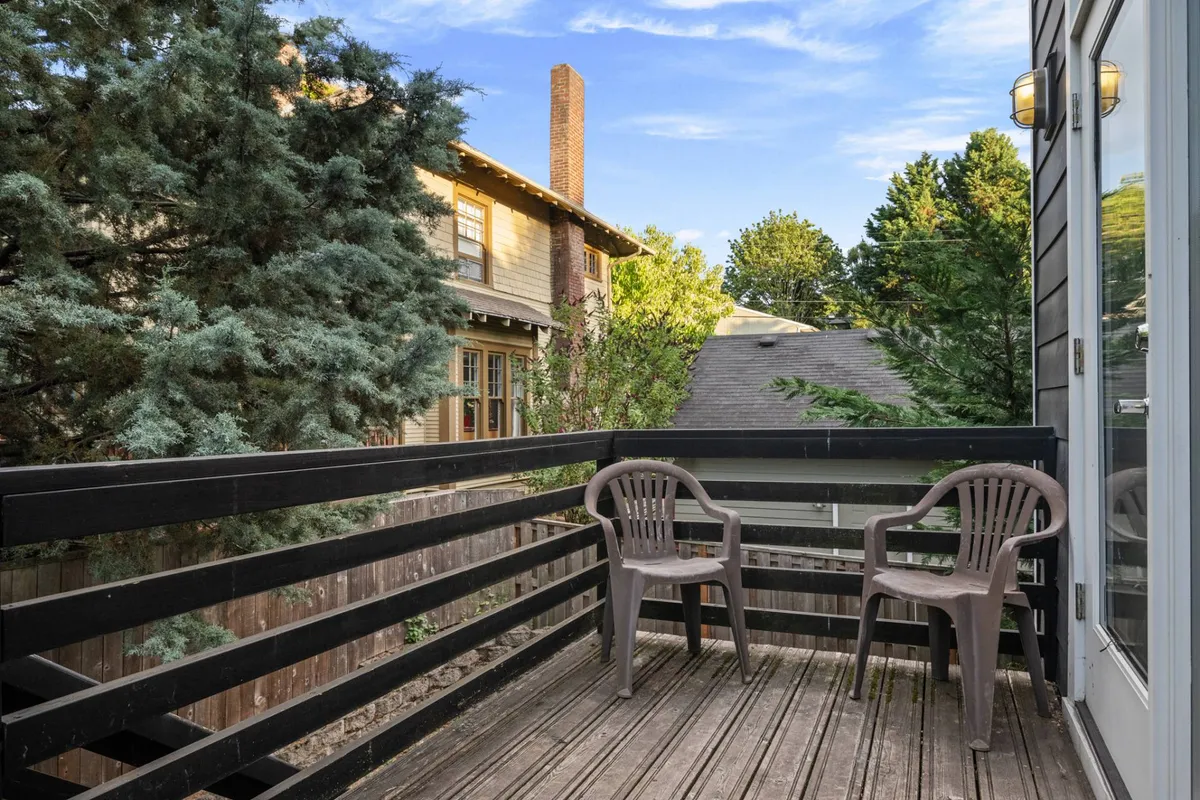 The image depicts an exterior wooden deck with two plastic chairs overlooking lush green trees in the background. The scene captures a relaxed, outdoor atmosphere ideal for enjoying morning coffee, reading, or simply soaking up the surrounding foliage.
