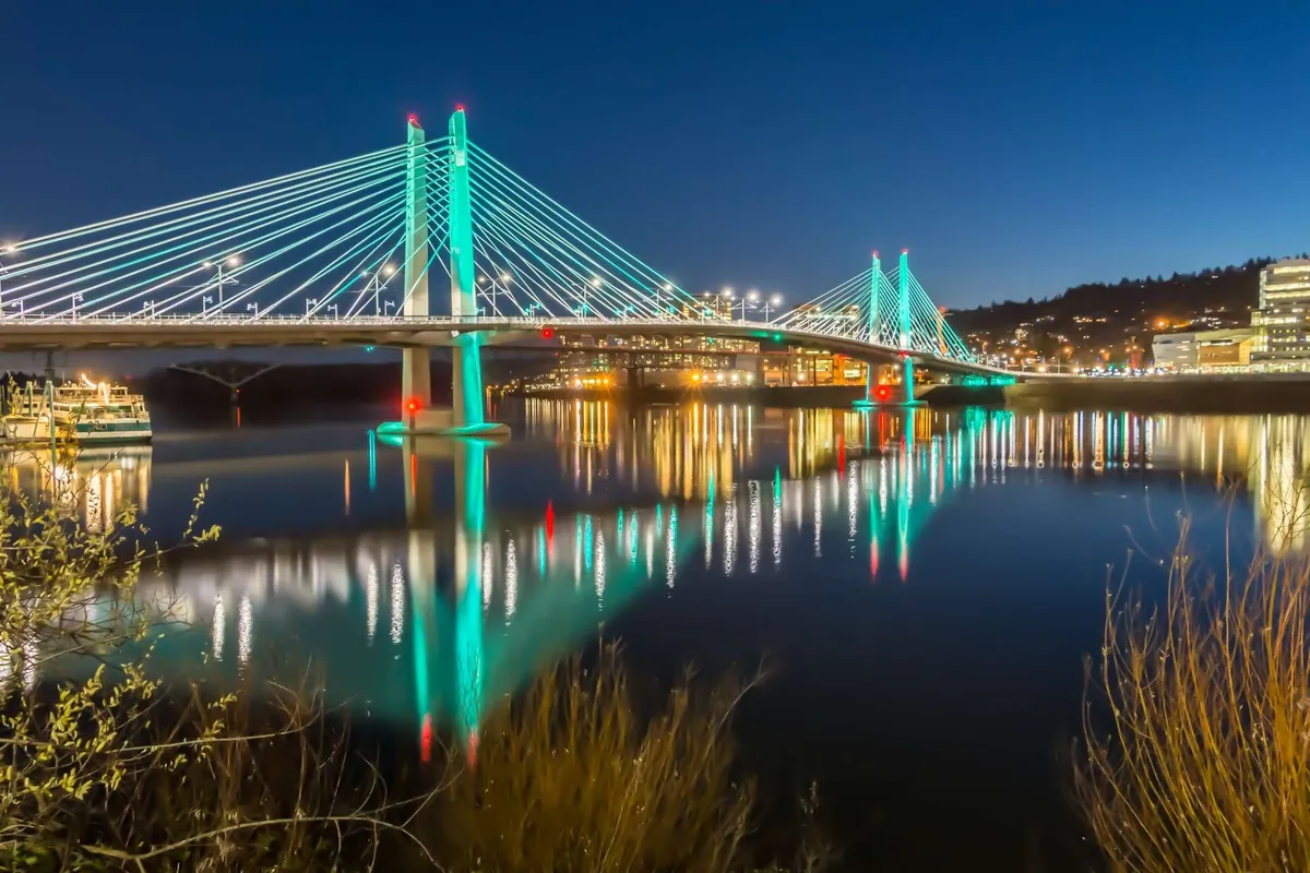 An evening view of an illuminated bridge over water with city lights reflecting in the background. The scene is lively and showcases the vibrancy of Portland's nearby attractions, hinting at adventures like the Eastbank Esplanade and other riverside activities.