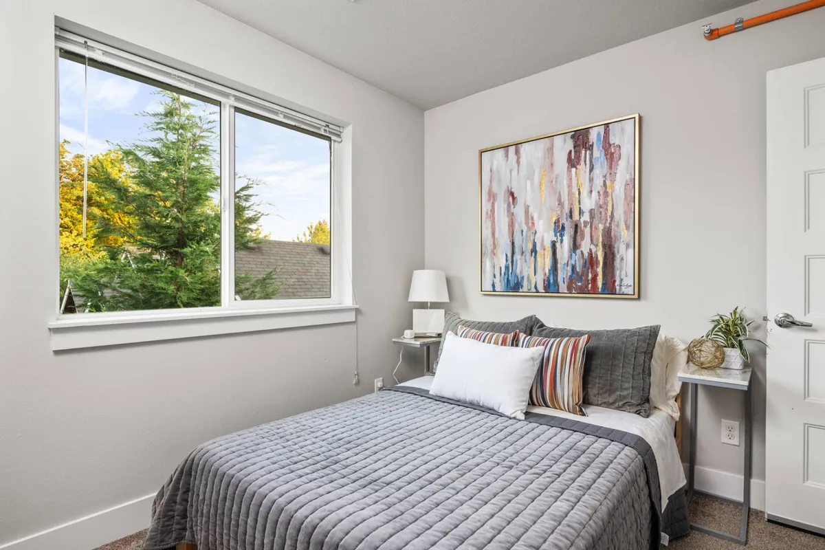 A tranquil bedroom with a bed dressed in a gray quilt, bathed in natural light from a nearby window. The space features a colorful abstract painting above the bed, a nightstand with a lamp, and an overall calm, restful atmosphere.
