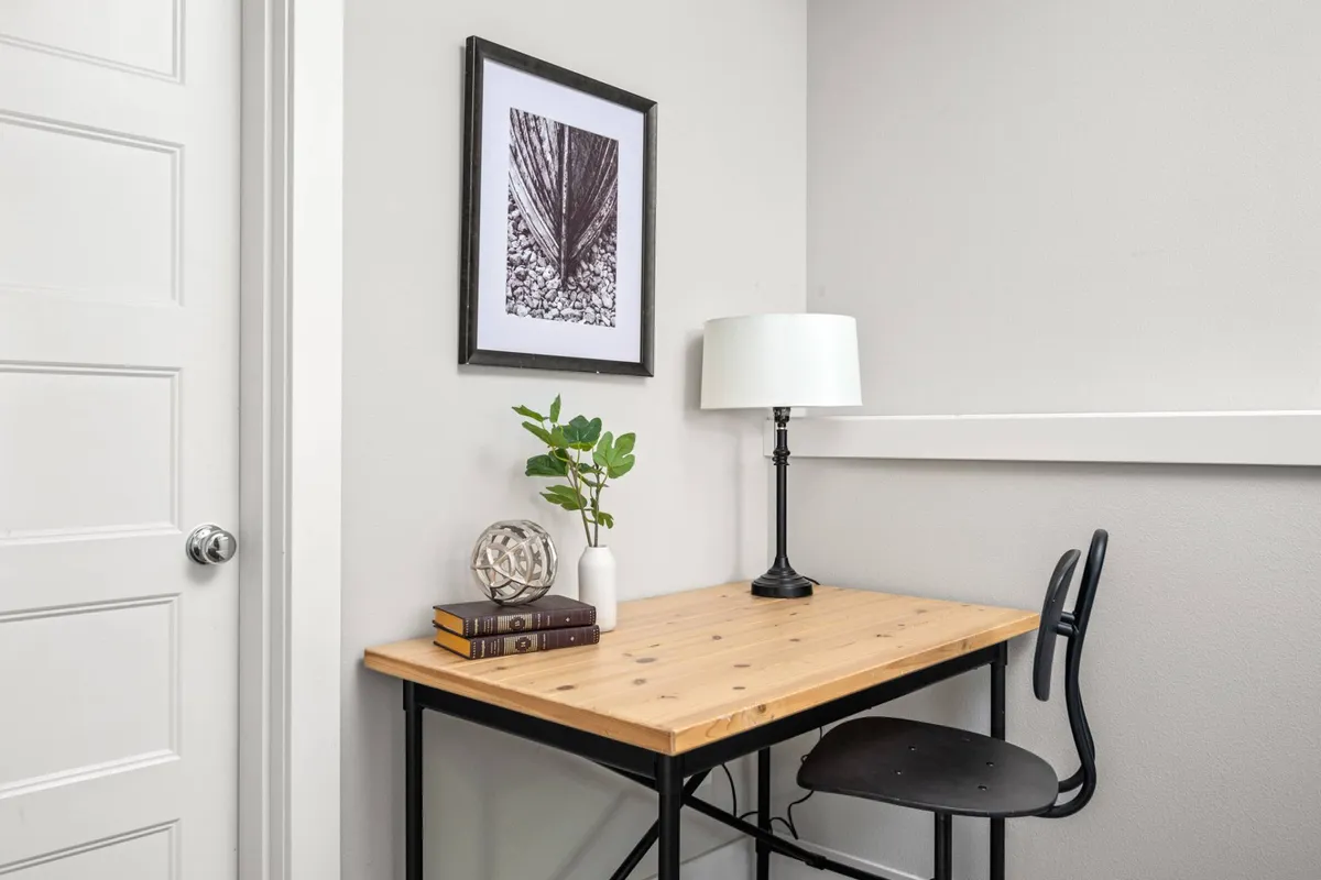 A tranquil workspace highlighted by a wooden table, desk lamp, framed artwork, potted plant, and a small stack of books. The mood is focused yet calm, ideal for reading, working, or studying.