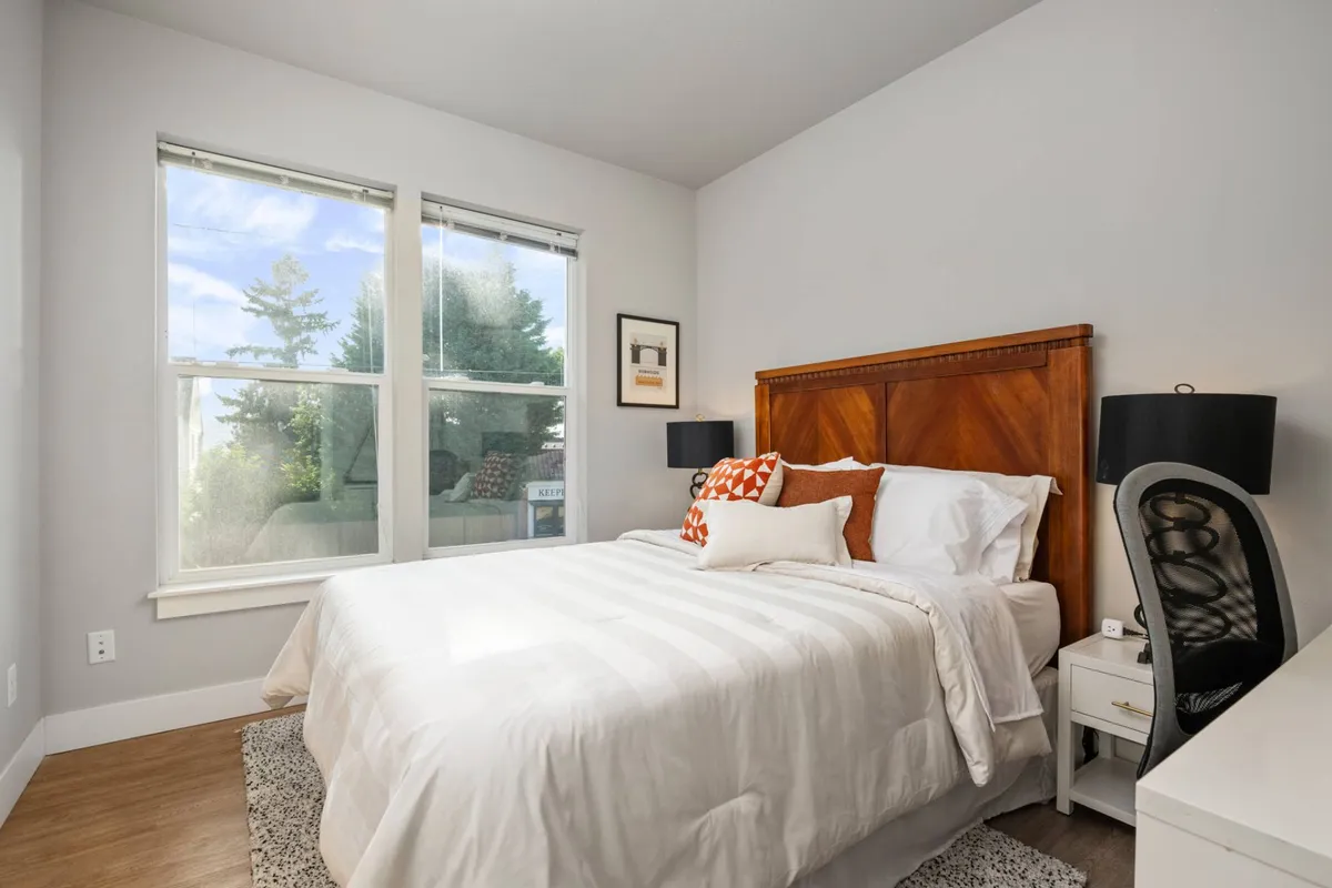 A peaceful bedroom with a bed, nightstand, decorative pillows, and windows letting in morning light. The mood is calm and inviting, with an emphasis on comfort and relaxation.