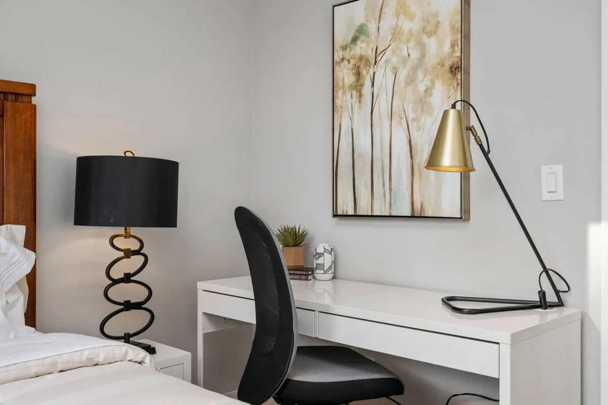 A serene bedroom featuring a white linen bed, a modern white desk with a black lamp, and artwork depicting trees. The space is both restful and conducive to productive work, supporting moments of quiet reflection or focused individual effort.