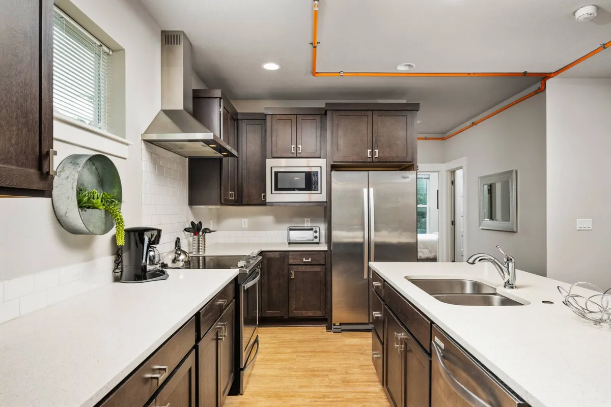 This image features an open, contemporary kitchen with stainless steel appliances, open shelving, a large countertop, and ample natural light streaming in through a window. The space is welcoming and designed for both cooking and socializing, making it ideal for informal team gatherings or meal prep together.