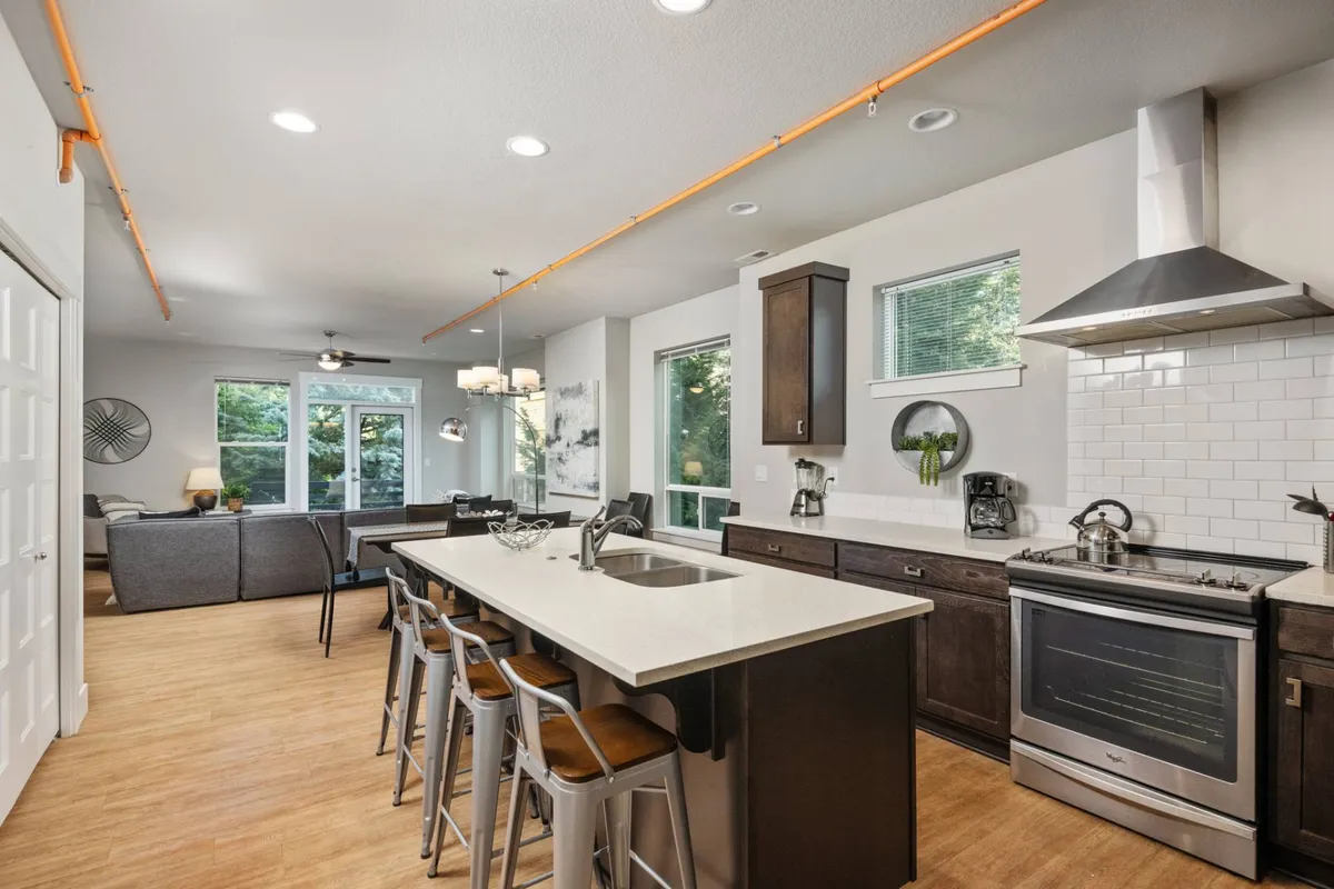 This image features a welcoming interior with a kitchen island, stainless steel appliances, and windows that overlook greenery. The open-plan space is bright, family-friendly, and designed for easy interaction among guests.