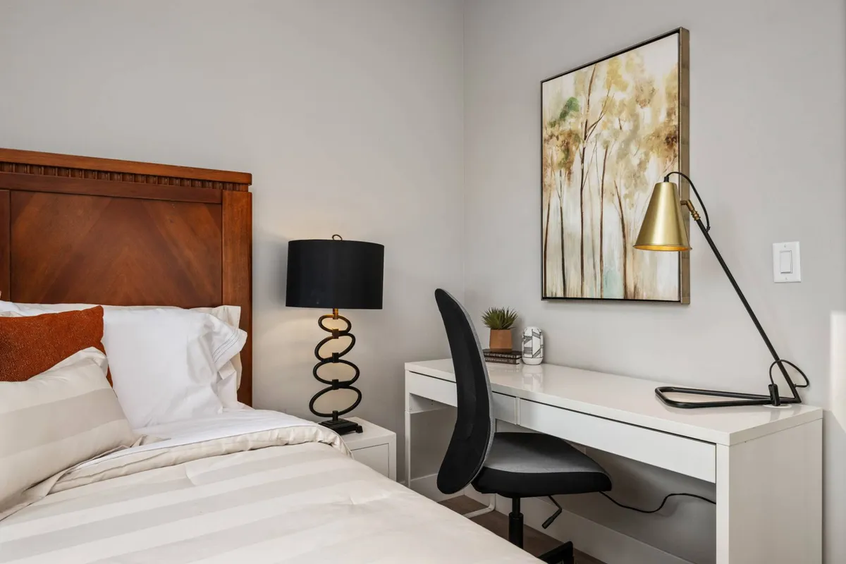 A cozy bedroom featuring a bed with striped bedding, a desk with a chair for work or reading, wall art, and a soft table lamp. The space is styled to allow for both relaxation and productivity, with an overall warm and inviting mood.