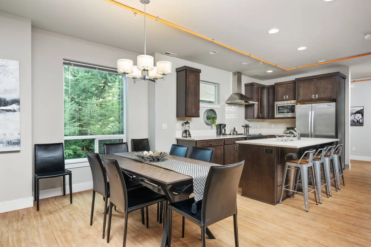 A bright, open-plan space featuring a spacious kitchen island, a large dining table set for a group, and wide windows letting in natural light. The scene is styled for family meals and entertaining, with elements that invite guests to gather for cooking, eating, and socializing.
