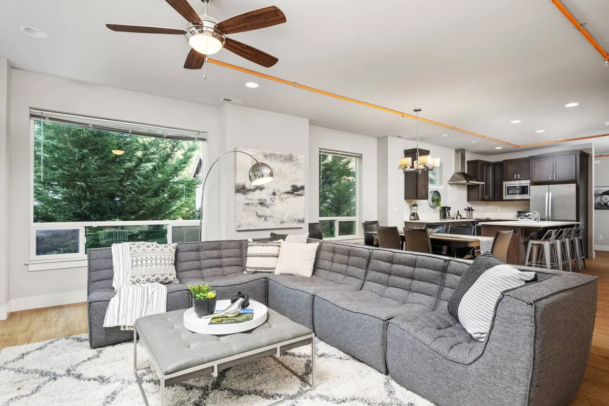 The image showcases a spacious, sun-filled living area featuring a large contemporary sectional sofa around a chic round coffee table, with a welcoming dining table in the background. Expansive windows frame lush greenery, infusing the space with natural light and offering tranquil views, creating a perfect environment for group gatherings, relaxation, or sharing meals.